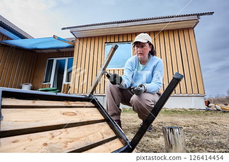 Applying anti-rust coating to metal parts of garden bench with brush, female house painter performs protective painting to prevent rust and damage. 126414454
