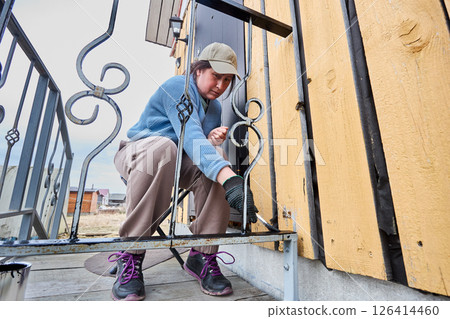 Applying anti corrosion compound to porch railing at entrance to rural house woman in her 50s uses paintbrush Applying anti corrosion compound to porch railing at entrance to rural house woman in her 50s uses paintbrush 126414460