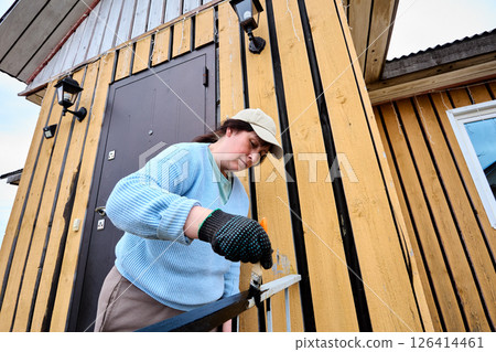 Painting metal railing with brush to protect surface from corrosion and improve appearance middle aged woman in cap works near entrance of wooden house. 126414461