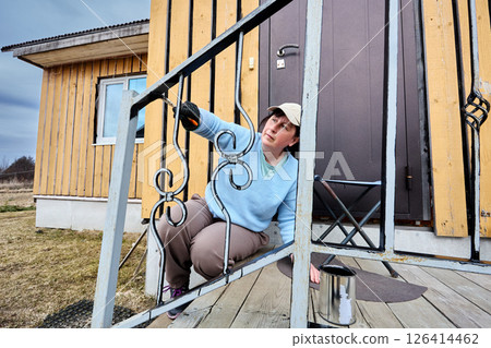 Applying black paint to metal railing of porch is performed by white woman over 50 sitting on step and leaning back with one arm extended forward. 126414462