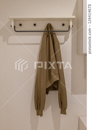 Wall mounted coat rack with white shelf and black hooks holding a khaki sweatshirt against white wall Wall mounted coat rack with white shelf and black hooks holding a khaki sweatshirt against white wall 126414678