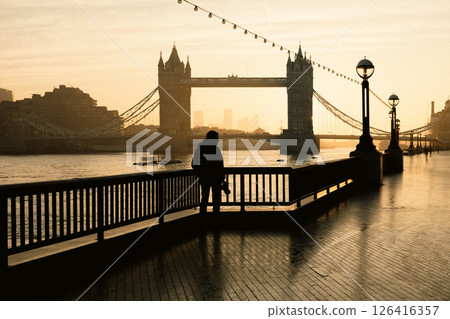 A photographer stands on Queen's Walk, admiring the tranquil morning view of Tower Bridge against a serene sunrise. The scene captures the essence of London waking up. A photographer stands on Queen's Walk, admiring the tranquil morning view of Tower Bridge against a serene sunrise. The scene captures the essence of London waking up. 126416357