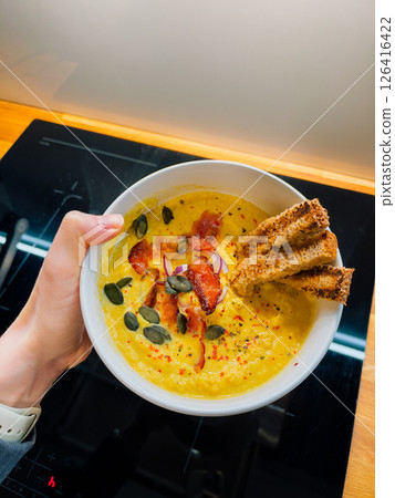 First-person view: hand holding bowl with pumpkin soup, bacon, seeds, toast, healthy textures ideal for wellness blogs, healthy lifestyle visuals for diet-friendly projects. Nutritious creamy soup POV 126416422