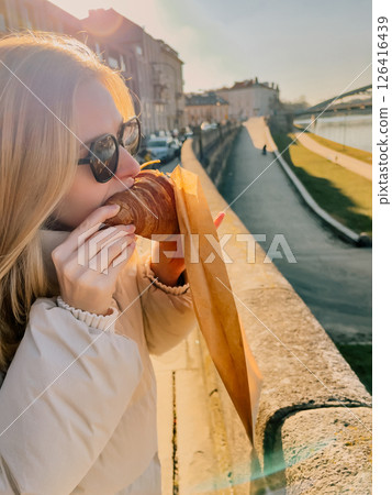 Blonde tourist wearing sunglasses and white jacket eating croissant from paper bag on bridge over Vistula river in Krakow, Poland, during sunny winter day. Blonde woman enjoys croissant from paper bag Blonde tourist wearing sunglasses and white jacket eating croissant from paper bag on bridge over Vistula river in Krakow, Poland, during sunny winter day. Blonde woman enjoys croissant from paper bag 126416439