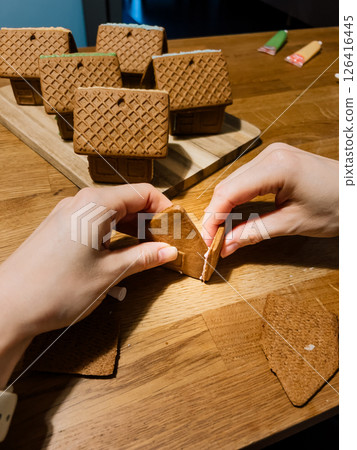 First-person view: Person carefully joining walls of gingerbread house with icing on wooden table, surrounded by other finished houses displayed on cutting board in background. Gingerbread house. POV First-person view: Person carefully joining walls of gingerbread house with icing on wooden table, surrounded by other finished houses displayed on cutting board in background. Gingerbread house. POV 126416445
