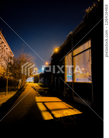 Empty sidewalk winding past industrial building cloaked in dry ivy, featuring lit window at night, casting elongated shadows on ground in tranquil darkness. Urban walkway, lluminated window, ivy 126416468