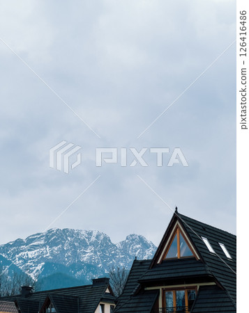Nestled in valley, traditional wooden houses with dark tiled roofs sit beneath dramatic, snow capped Tatra Mountains, creating serene winter landscape. Wooden house with dark roof in mountain valley 126416486
