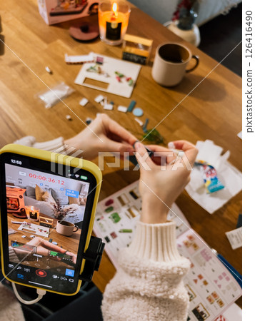Video shows woman crafting at table, video setup with tripod and phone, video captures step-by-step work, video records creative DIY moment. Tutorial filmed in cozy home, hands prepare craft tutorial Video shows woman crafting at table, video setup with tripod and phone, video captures step-by-step work, video records creative DIY moment. Tutorial filmed in cozy home, hands prepare craft tutorial 126416490