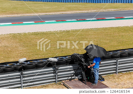 Cameraman crew stands large pro video camera on motorsport race track shaded heat black umbrella hot summer day. TV equipment safety tires fence automotive moto challenge auto racing Cameraman crew stands large pro video camera on motorsport race track shaded heat black umbrella hot summer day. TV equipment safety tires fence automotive moto challenge auto racing 126416534