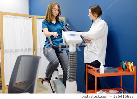 Female doctor cardiologist measures pressure with patient during bicycle exercise for examination cardiovascular system at medical office of clinic. Young woman training on bike simulator. 126416652