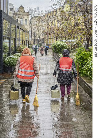 Women wearing safety vests cleaning wet city street during light snowfall. Bad weather, climate change, mud and puddles. Concept of outdoor public service, city maintenance 126416670