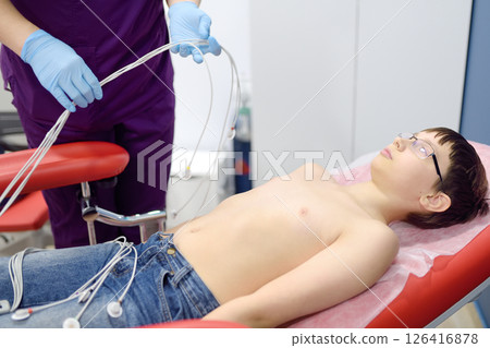 Child under ECG test with doctor. Boy patient lying in medical bed with vacuum sensors. A nurse performs an electrocardiogram of the heart in office of a medical clinic. 126416878