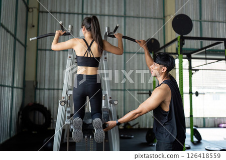 Wellness and Fitness. Personal trainer assisting a woman with pull-up exercise in gym. Wellness and Fitness. Personal trainer assisting a woman with pull-up exercise in gym. 126418559