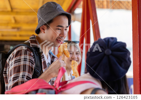 Young Asian man engaging in a lively conversation with friends while enjoying street food at a vibrant market, emphasizing travel and friendship. Young Asian man engaging in a lively conversation with friends while enjoying street food at a vibrant market, emphasizing travel and friendship. 126423105