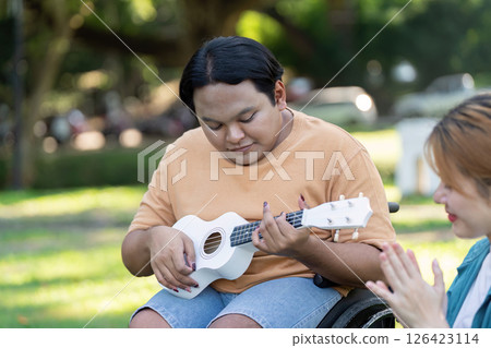 Young man in a wheelchair playing ukulele while a friend claps in appreciation. Young man in a wheelchair playing ukulele while a friend claps in appreciation. 126423114
