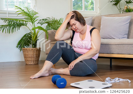 Overweight woman sitting on the floor, feeling overwhelmed on her weight loss journey next to dumbbells and a scale Overweight woman sitting on the floor, feeling overwhelmed on her weight loss journey next to dumbbells and a scale 126425620