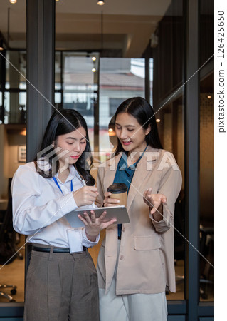 Collaborative women discussing project details while reviewing data on digital tablet in modern office Collaborative women discussing project details while reviewing data on digital tablet in modern office 126425650