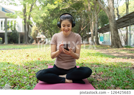 Wellness and Music Routine. A woman wearing headphones, enjoying music while engaging with her phone in a tranquil park setting. 126425683