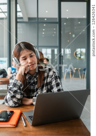 A young woman wearing headphones, focused on her laptop in a contemporary cafe environment. 126426341