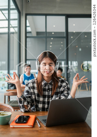 Young woman in plaid shirt meditating with headphones at a laptop in a modern office setting, promoting mindfulness and productivity. 126426344