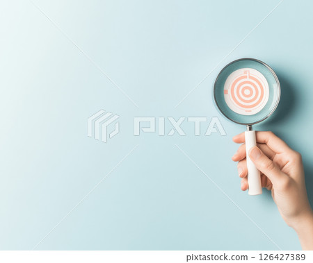 Hand holding magnifying glass over target symbol on blue background, representing focus and clarity in business objectives Hand holding magnifying glass over target symbol on blue background, representing focus and clarity in business objectives 126427389