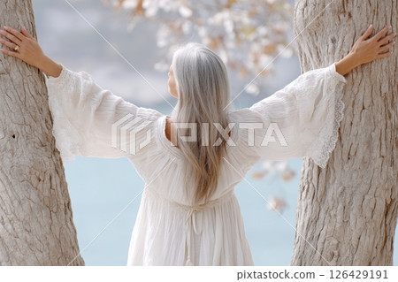 Woman in flowing white dress standing between two trees with arms outstretched, facing serene blue water in peaceful natural setting 126429191