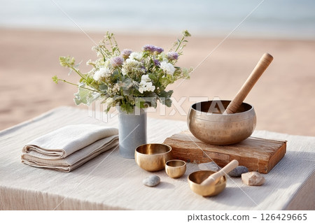 Still life of singing bowls, flowers and folded linens on a table by the beach, evoking calm and wellness 126429665