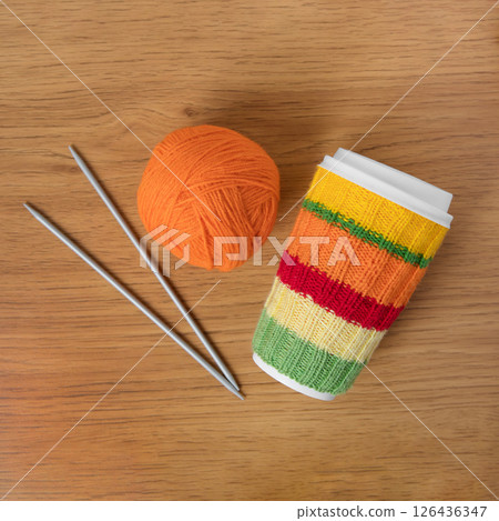 Cup for takeaway coffee with knitted autumn striped cover and knitted wool with knitting needles on wooden background. Concept of takeaway coffee, walking, energetic life, hobbies needlework Cup for takeaway coffee with knitted autumn striped cover and knitted wool with knitting needles on wooden background. Concept of takeaway coffee, walking, energetic life, hobbies needlework 126436347