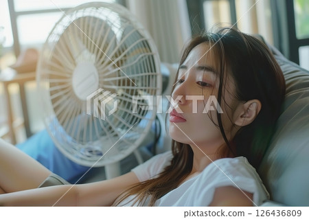 Young woman suffering during a heat heat sitting under air conditioner at home resting on a sofa. Exhausted girl feeling sweaty switching on modern AC system. Hot summer. Fresh clean air in apartment 126436809