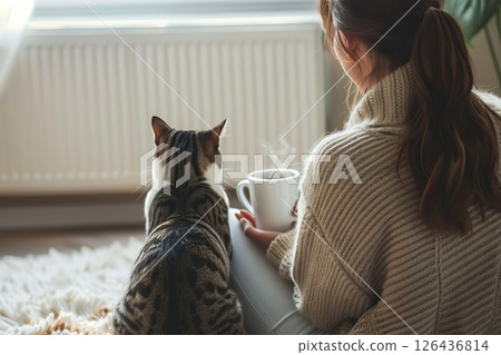 Young woman wearing a sweater freezing for winter cold with her cat. Girl is sitting over electric heater, drinking hot coffee or tea, warming up hands. Discomfort spending time at home. Coldly, chill 126436814