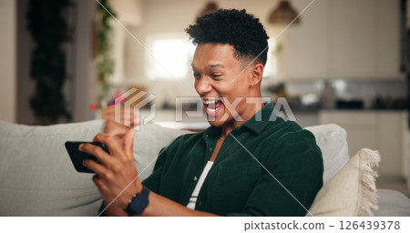 Gaming, phone and fist pump with man on sofa for esports streaming, cyber sport league and winner. Celebration, gamer success and app platform with excited person in home for online tournament 126439378