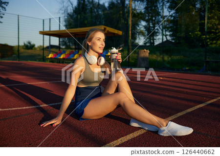 Dreamy relaxed sportswoman drinking water sitting on basketball court Dreamy relaxed sportswoman drinking water sitting on basketball court 126440662