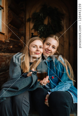 Two women sitting closely on steps of rustic log cabin, sharing hot drink and enjoying each other's company while embracing warmth of chilly autumn day. Two girl friend sitting together on porch 126441159