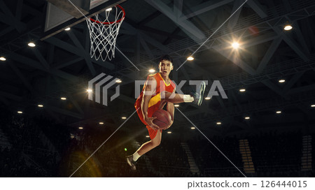 Conceptual collage. Basketball player jumps high holds ball under basket on 3D arena with lights and visible metal roof structure. Conceptual collage. Basketball player jumps high holds ball under basket on 3D arena with lights and visible metal roof structure. 126444015