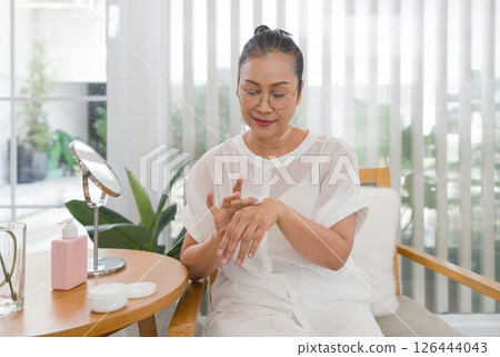 Senior asian woman sit at a wooden table, applying a skincare product to her hand. She enjoy bright natural light filtering through sheer curtains in a cozy home environment. Senior asian woman sit at a wooden table, applying a skincare product to her hand. She enjoy bright natural light filtering through sheer curtains in a cozy home environment. 126444043
