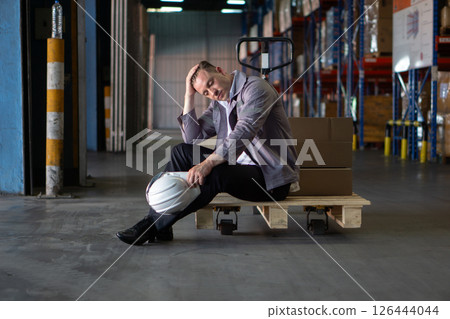 A warehouse worker rest on a wooden pallet surrounded by cardboard boxes. He appear exhausted, with a hard hat in hand, reflecting on a busy workday. 126444044