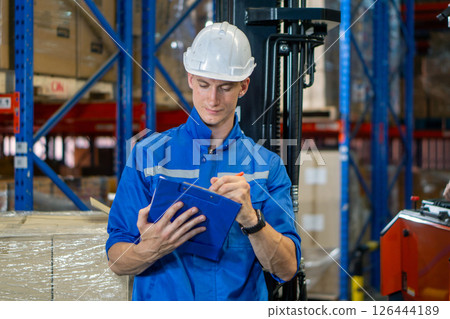 Young worker in blue uniform and safety helmet review inventory record on clipboard. Stand among stacked boxes in a busy storage area. Working atmosphere in general warehouse with racking system Young worker in blue uniform and safety helmet review inventory record on clipboard. Stand among stacked boxes in a busy storage area. Working atmosphere in general warehouse with racking system 126444189