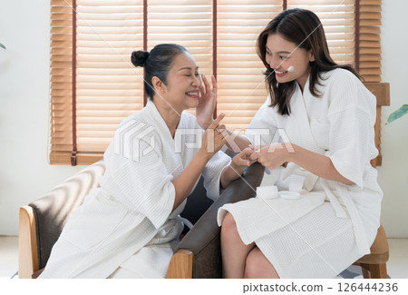 Asian mother and daughter in white bathrobe enjoy a pampering experience at home. They engage in skincare routine while sharing smile and laughter, creating a joyful atmosphere. Asian mother and daughter in white bathrobe enjoy a pampering experience at home. They engage in skincare routine while sharing smile and laughter, creating a joyful atmosphere. 126444236