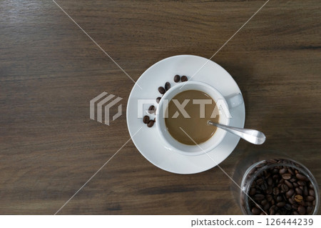 A white cup filled with hot coffee sits on a saucer next to several roasted coffee bean. A glass jar with more coffee beans is visible nearby, creating a cozy morning atmosphere. 126444239