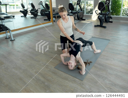 In a contemporary gym setting, an instructor provide guidance to a student performing yoga bridge pose. Sunlight stream through large window, enhancing the focused atmosphere. In a contemporary gym setting, an instructor provide guidance to a student performing yoga bridge pose. Sunlight stream through large window, enhancing the focused atmosphere. 126444491