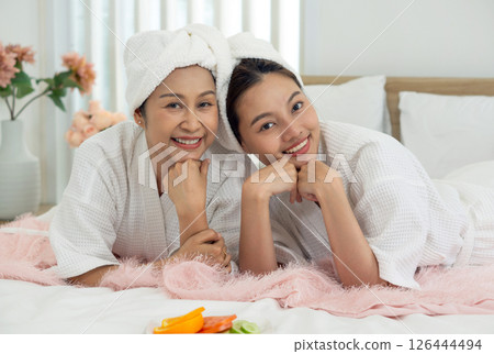 Two asian women, smiling and wearing white bathrobe, relax on a plush bed in a bright room decorated with flower. They enjoy each other's company during a wellness getaway. Two asian women, smiling and wearing white bathrobe, relax on a plush bed in a bright room decorated with flower. They enjoy each other's company during a wellness getaway. 126444494