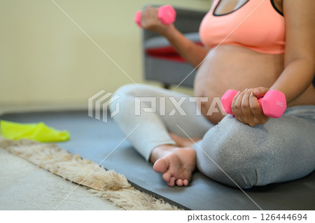 Close up of pregnant woman sitting holding pink dumbbells for a light home workout Close up of pregnant woman sitting holding pink dumbbells for a light home workout 126444694