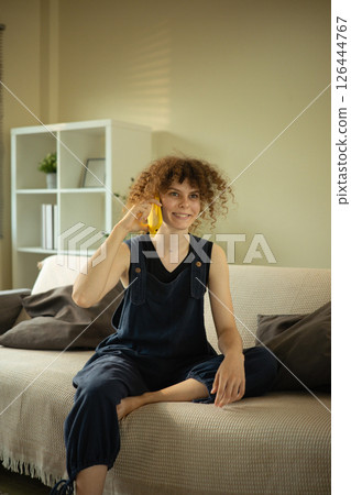 Happy young woman with curly hair talking on the phone sitting in bright living room Happy young woman with curly hair talking on the phone sitting in bright living room 126444767