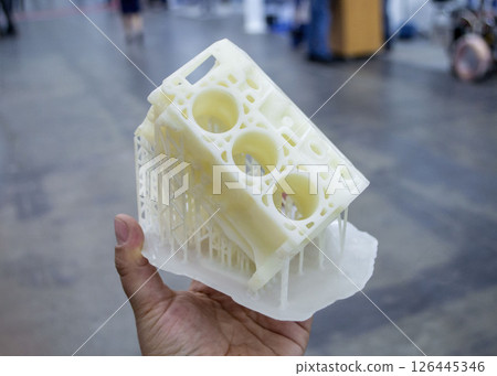 Person holding in palm of hand prototype of car engine printed on 3D printer from molten white plastic. Three-dimensional model. Modern new printing industry. Additive progressive modeling technology. Person holding in palm of hand prototype of car engine printed on 3D printer from molten white plastic. Three-dimensional model. Modern new printing industry. Additive progressive modeling technology. 126445346