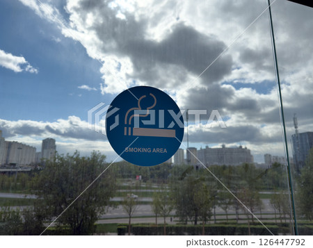 Blue smoking area sign on glass wall with urban view in background, illustrating the widespread normalization of smoking and growing demand for designated spaces in modern public environments Blue smoking area sign on glass wall with urban view in background, illustrating the widespread normalization of smoking and growing demand for designated spaces in modern public environments 126447792