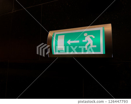 Illuminated green emergency exit sign on dark wall with arrow pointing left, symbolizing clear safety guidance and effective evacuation routes in case of fire or other critical situations 126447793