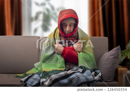 Middle age male model in hat parka and plaid getting cold sitting on the sofa in cold living room 126450354