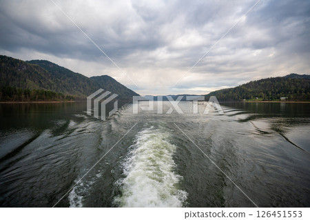 Wake trail behind ferry moving across Teletskoye lake in Altai Russia. Tranquil water surface with distant shoreline and mountains Wake trail behind ferry moving across Teletskoye lake in Altai Russia. Tranquil water surface with distant shoreline and mountains 126451553