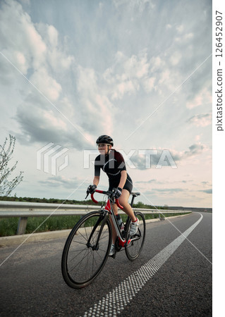 Confident girl, cyclist in full gear riding along open countryside road, powerful stride and focused eyes locked on route ahead. Confident girl, cyclist in full gear riding along open countryside road, powerful stride and focused eyes locked on route ahead. 126452907