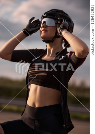 Smiling female athlete adjusting reflective helmet and sunglasses while enjoying warm sunset light after bike ride, intense training outdoor. 126452925